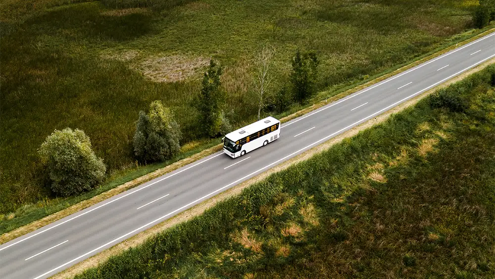 Vista aérea de un autobús blanco circulando por una carretera entre zonas verdes de campo, en un entorno rural despejado.