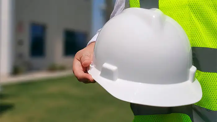 Persona con chaleco reflectante sosteniendo un casco de seguridad blanco en la mano, en un entorno exterior con fondo desenfocado.