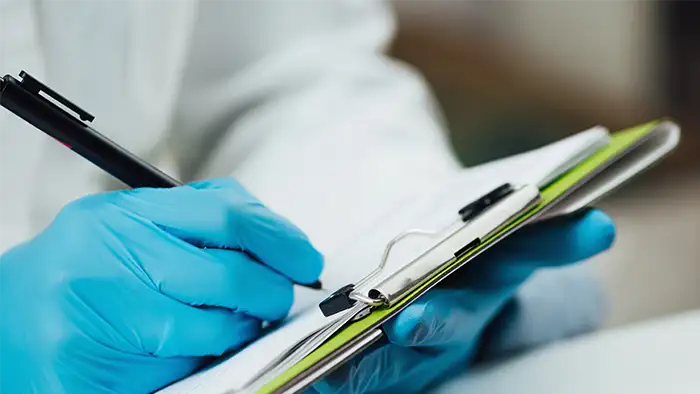 Mano con guante de nitrilo azul escribiendo con un bolígrafo sobre un portapapeles con documentos, en un entorno de trabajo técnico o sanitario.